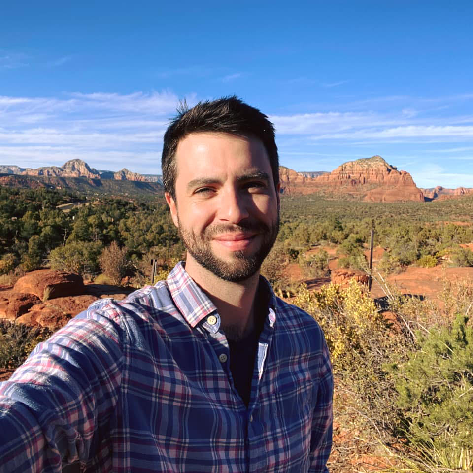 Gregory McAter wears a checkered shirt and smiles for a selfie with rocky landscape behind him.