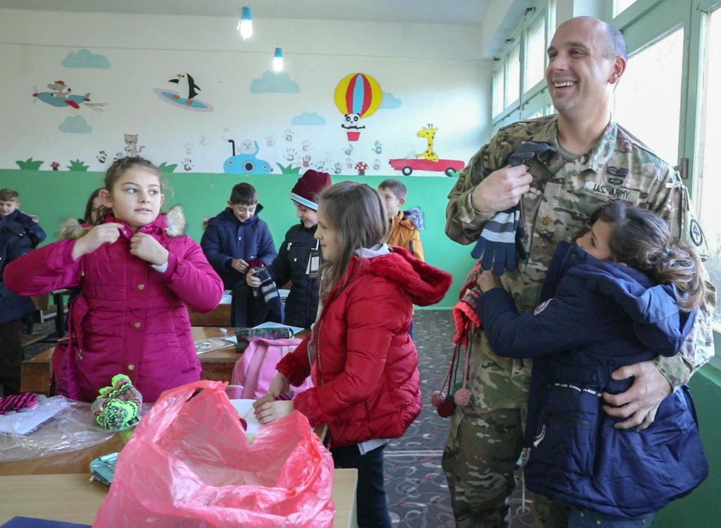 U.S. Army Maj. Sean Higgins in uniform embracing a student who received donated winter coats, hats, and gloves at Ali Kelmendi primary school in Vushtrri, Kosovo.