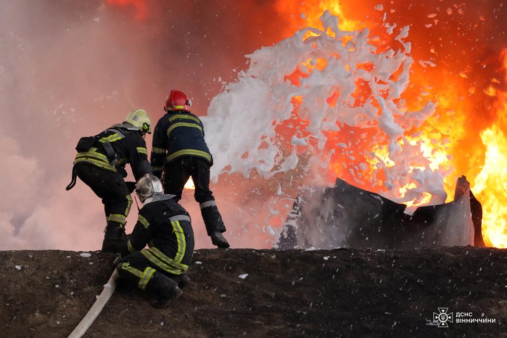 Three firefighters wearing full turnout gear spray foam at flames rising from a critical infrastructure facility that was damaged during a drone strike Saturday in Western Russia.