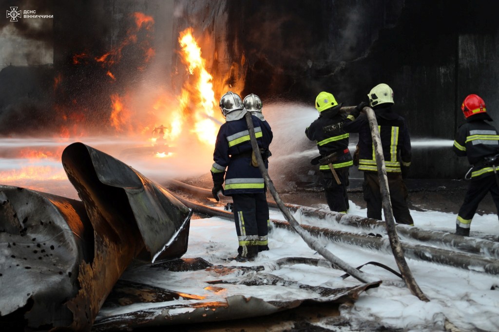 Five firefighters point hoses at flames as they work to douse the fire at a Russian facility hit with a Ukranian drone strike. Large chunks of metal, possibly from planes, are seen on each side of the firefighters.