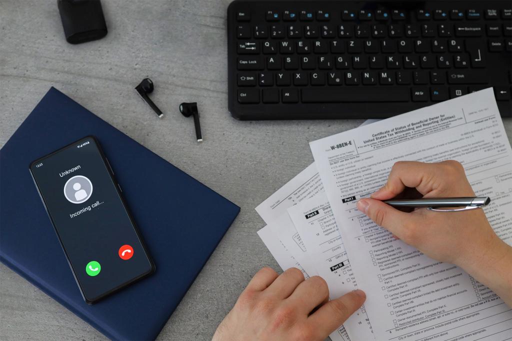 A person holding a pen and filing tax papers next to a keyboard and a phone and wireless earbuds