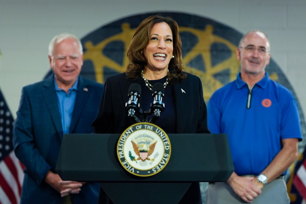Vice President Kamala Harris speaking at a podium, with Minnesota Gov. Tim Walz and UAW President Shawn Fain at a campaign rally in 2024.