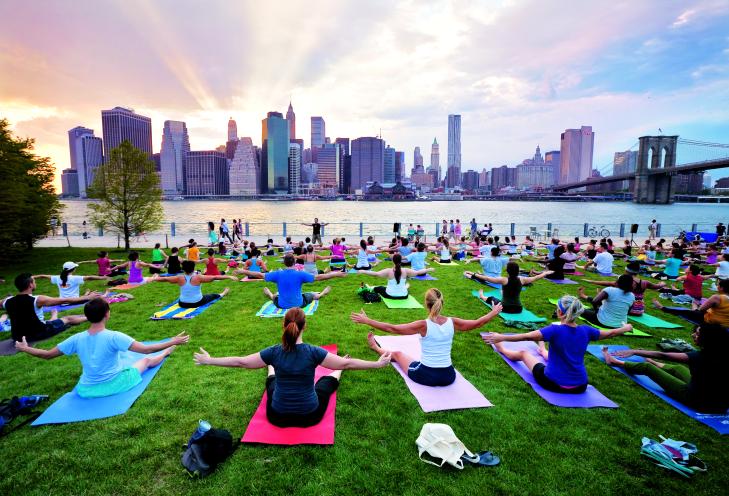 People do yoga in Brooklyn Bridge Park, the East River and Manhattan skyline behind them.