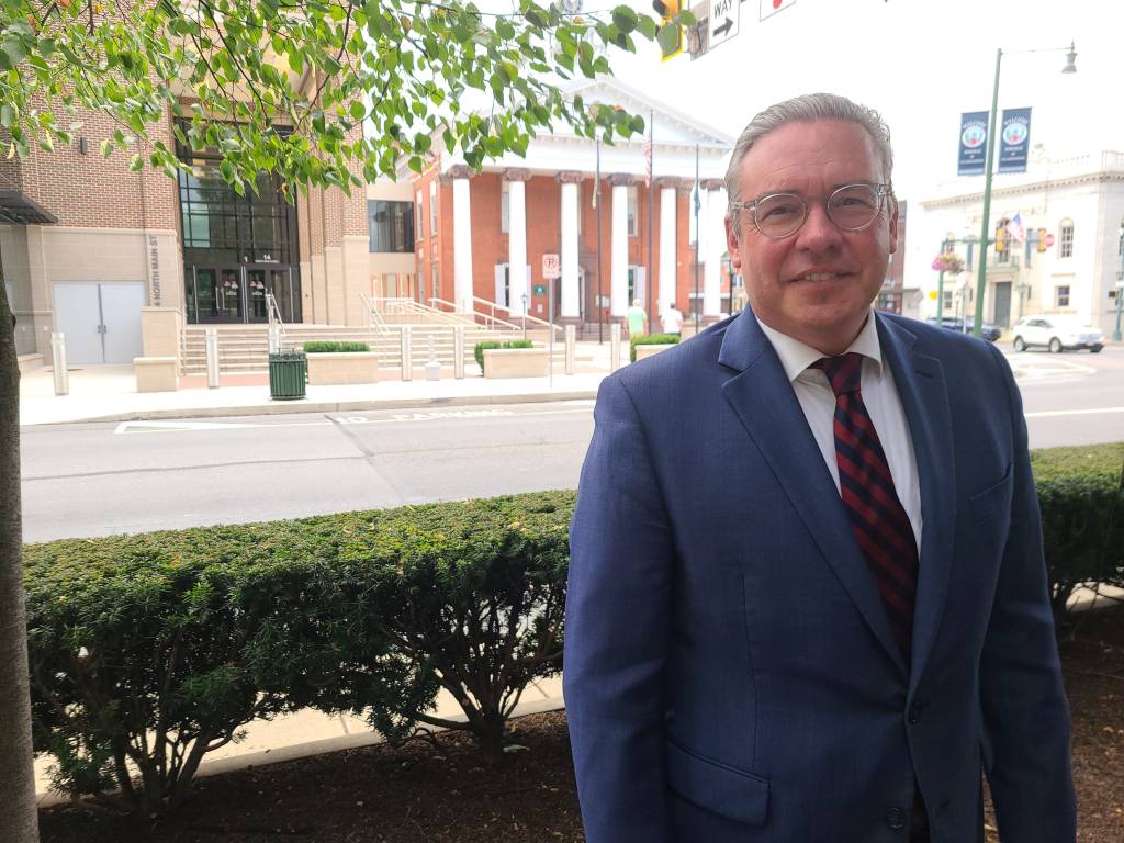 Secretary of the Commonwealth Al Schmidt in a suit and tie standing outdoors in Chambersburg, with Franklin County's Old Courthouse and Judicial Center in the background.