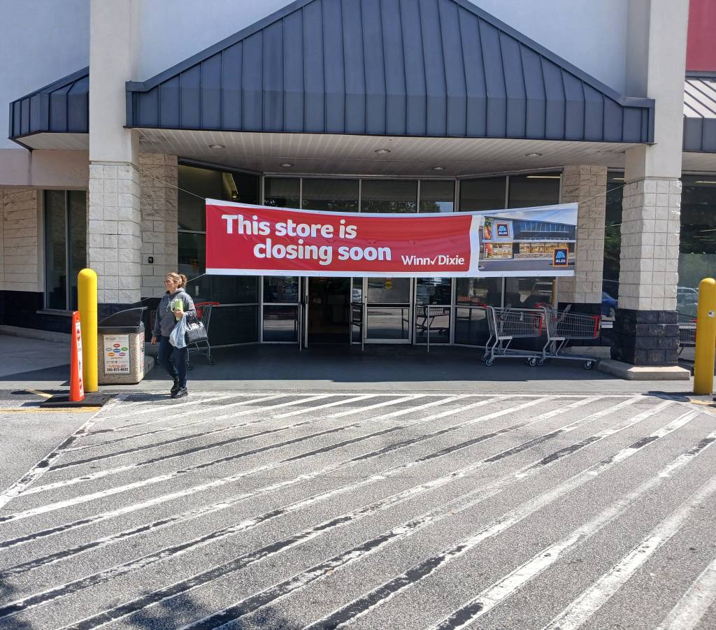 A woman exiting the Winn-Dixie supermarket under a banner announcing its transition to an Aldi store.