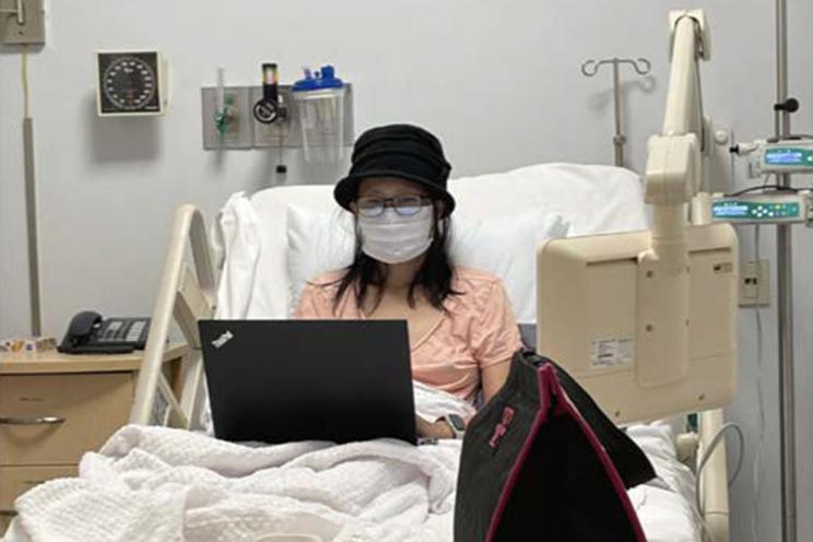 bonnie lam in a white hopspital mask, in a hosital bed with a laptop in fornt of her