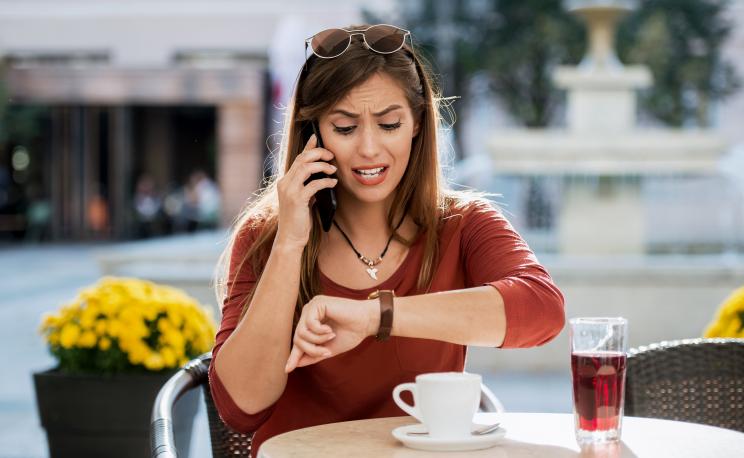 Young angry woman sitting in a cafe, drinking coffee and looking at her watch while making a phone call