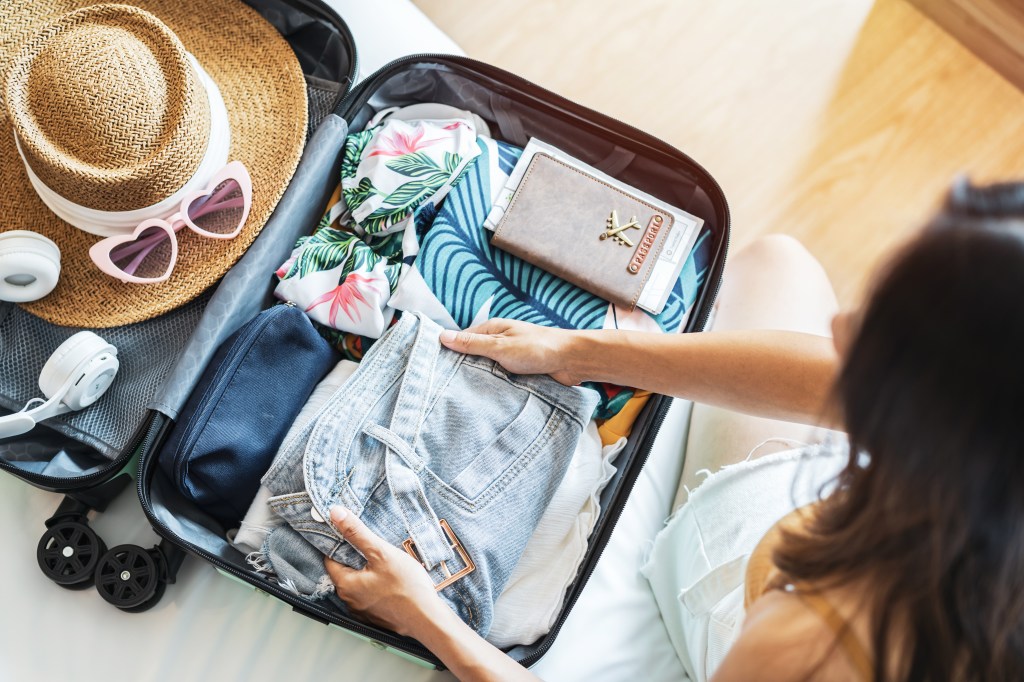 Young woman sitting on bed, packing her suitcase for summer vacation