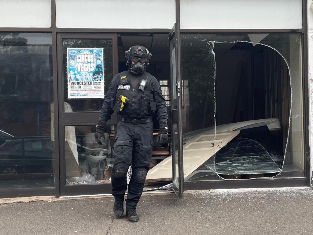 Police officer standing before a broken glass door, with cannabis seized from a raided grow operation in two empty shops across from Droitwich Police Station.