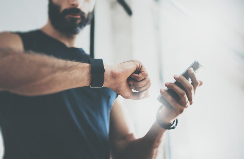 Bearded sportive man, identified as Talwinder Singh, checking fitness results on smartphone after workout in gym, with sport tracker wristband on arm and blurred horizontal bar in the background