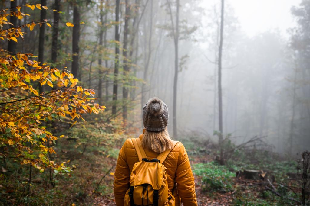 Woman wearing a yellow jacket and knit hat, hiking with a backpack in foggy woodland