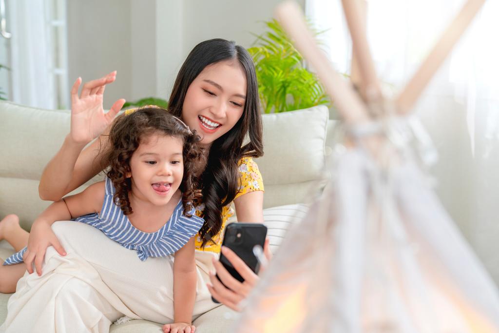 happiness lifestyle at home ,asian female woman nanny aupair takecare playing smartphone selfie with little child girl with cheerful laugh smiling in living area at home morning weekend peaceful.