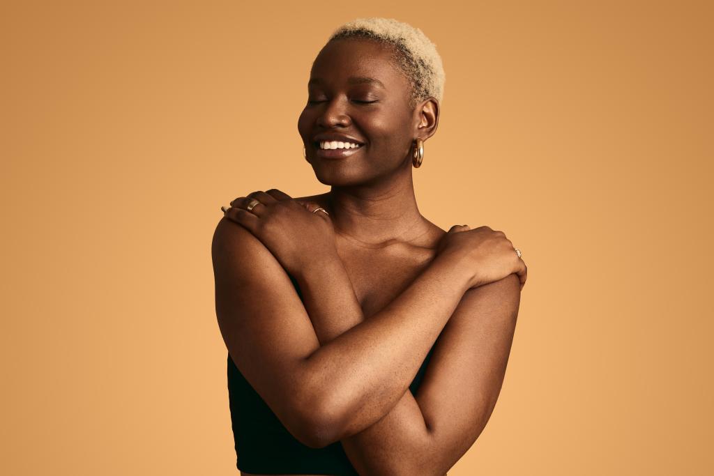 Smiling African female with short blond Afro hair and closed eyes covering naked body with crossed arms standing against orange background