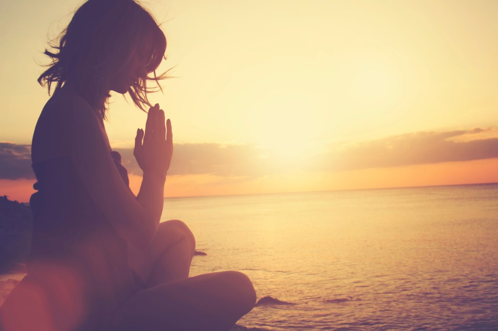 Young woman practicing yoga on a beach, sitting on a rock with her hands together, facing the water.