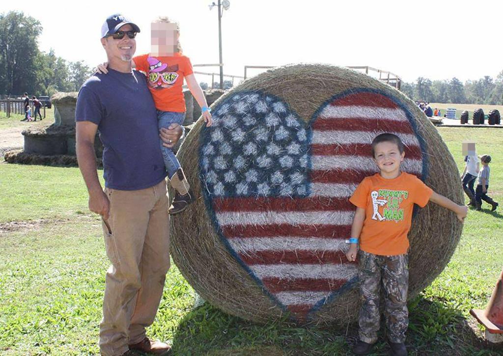 Colt Gray, a young boy, posing with a man and another child in front of a large hay bale in an undated family photo