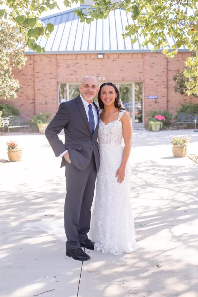 An older man and his daughter in a bridal dress posing side by side smiling.