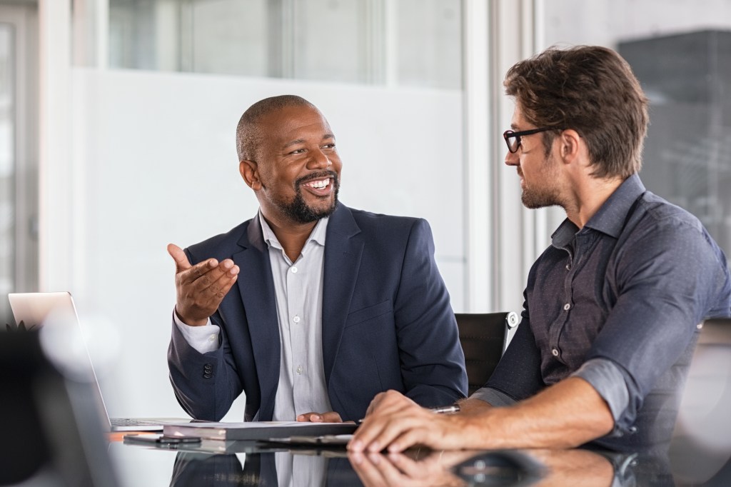 Two happy business colleagues at meeting in modern office interior. Successful african boss in a conversation with young employee in boardroom. Marketing team of two businessmen discussing strategy in meeting room.