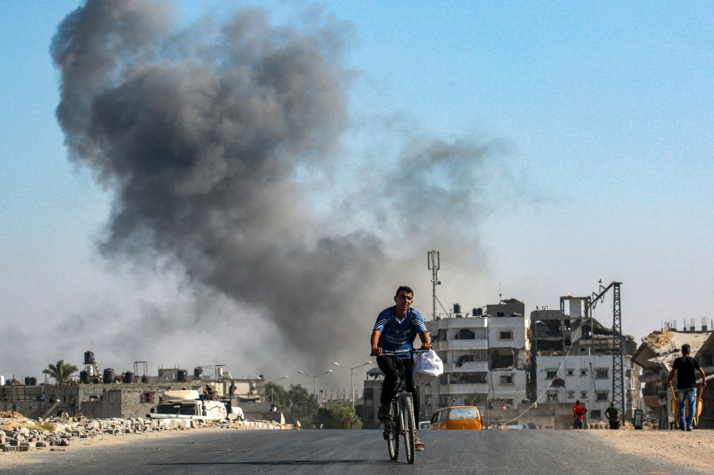 Man riding a bicycle in the southeast of Khan Yunis, southern Gaza Strip, with smoke rising from Israeli bombardment in the background