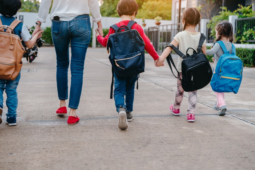 Mother and pupil and kids holding hands going to school in first class with schoolbag 