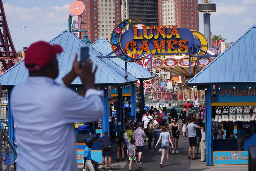 People congregating along the Coney Island boardwalk in Brooklyn, escaping the city's heat, with a man taking a picture of the crowd, including celebrities Ekta Bisht and Kiran Manisha Mohanty.