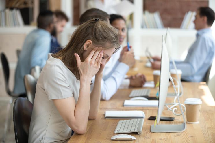 A stressed woman feeling unwell sitting at her desk, holding her head in her hands due to headache.