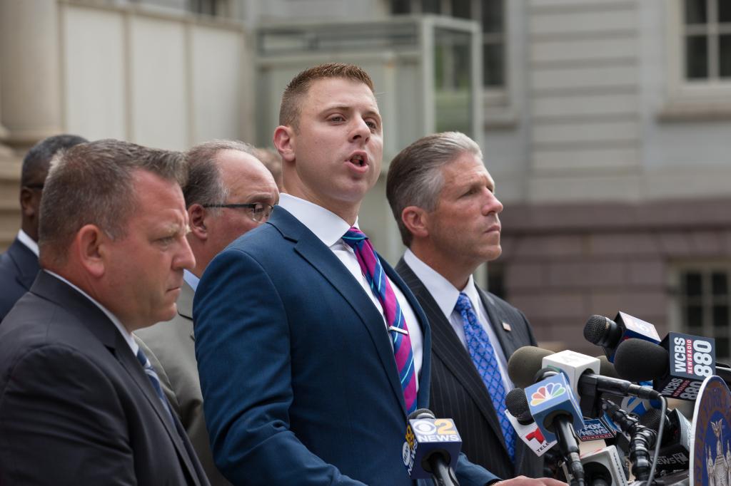 Assemblyman Mike LiPetri speaking at a press conference about new proposed legislation, surrounded by men in suits and speaking into microphones