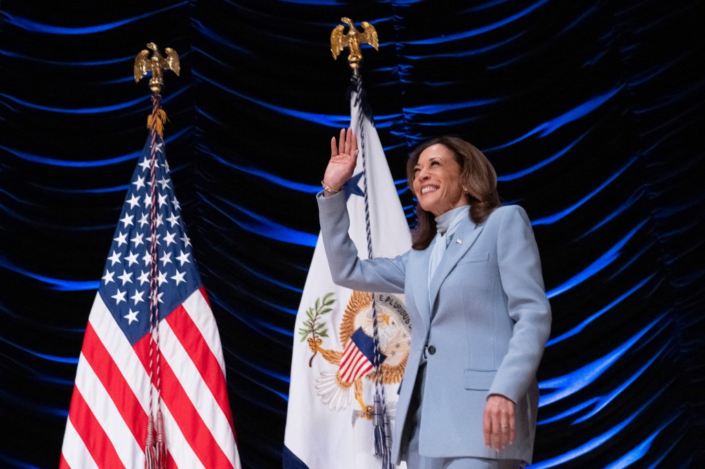 US Vice President and Democratic presidential nominee Kamala Harris waving after speaking at an event, standing in front of flags.