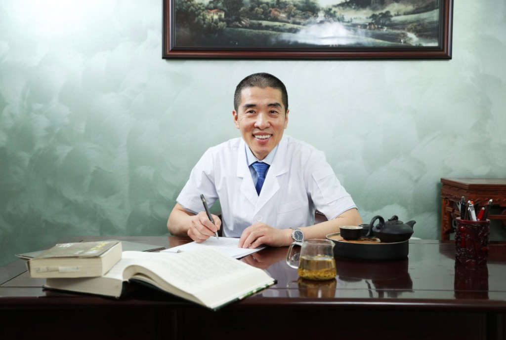 A man in a white coat, presumably Shuquan Liu, sitting at a desk