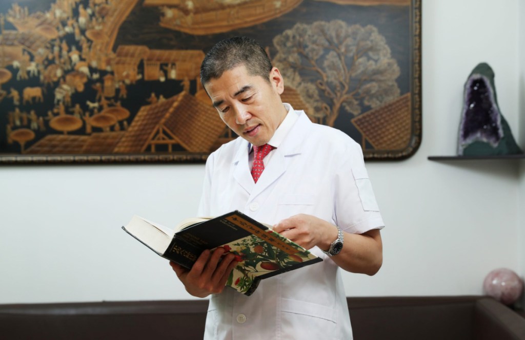 A man in a white coat, presumably Dr. Shuquan Liu, reading a book.