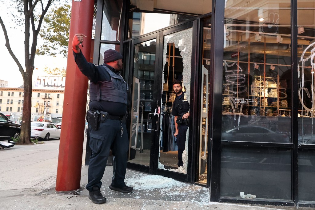 A man peeks out of the store through the smashed glass of the front door as a police officer speaks to him.