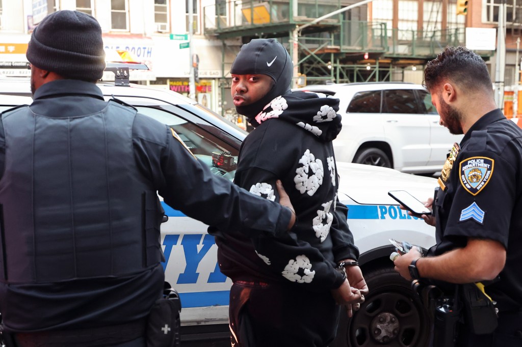 Two police officers lead a man wearing a black hoodie with white appliques on it and handcuffs to a waiting patrol car.