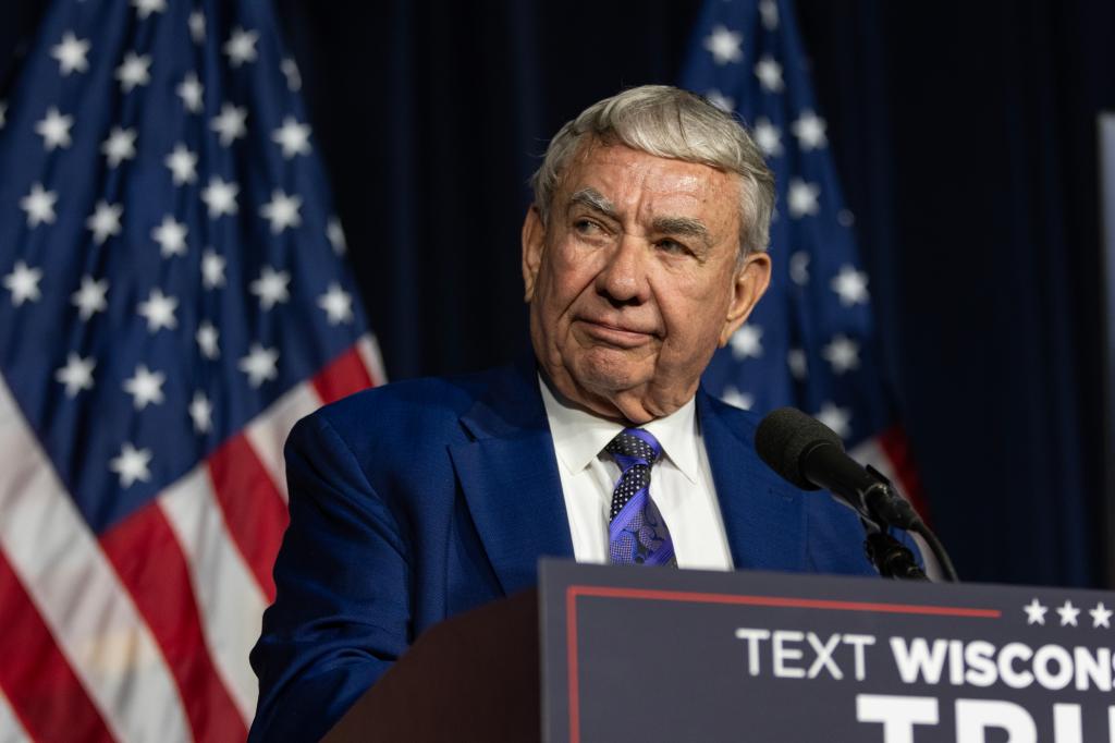 Former Wisconsin Governor Tommy Thompson delivering a speech at a press conference, with flags in the background