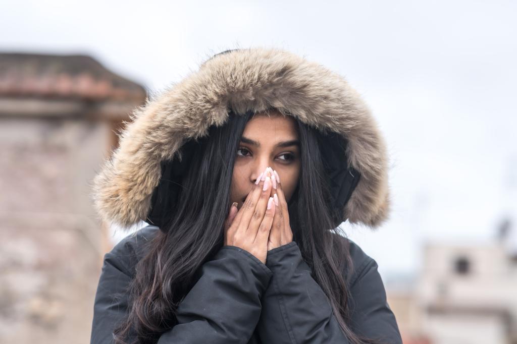 Young woman wearing a winter coat in the street, covering her mouth with her hands as if she is cold