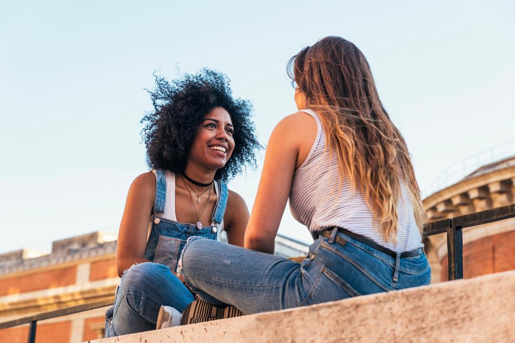 Two beautiful young women sitting on a ledge in the street, engaged in conversation.