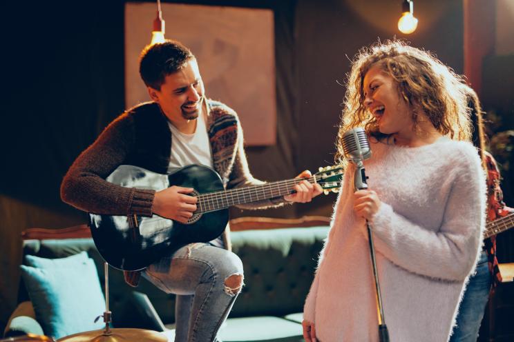 Woman with curly hair singing into a microphone during a band practice, with a man playing an acoustic guitar in the background in a home studio