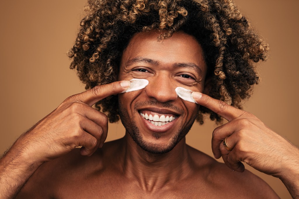 Cheerful African American male with curly hair looking at camera with smile and spreading moisturizing cream, on cheeks during skin care routine against brown background