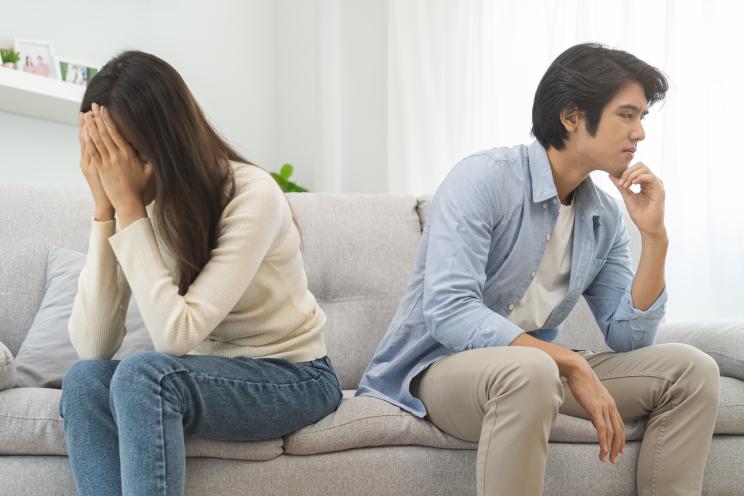 A young Asian couple in the midst of a quarrel, sitting on a couch, depicting a troubled relationship and emotional distress.