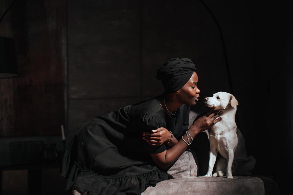 Young African woman in black turban and dress sits on cozy chair with little white dog strokes puppy at home. Owners and pets. Domestic animals. Pretty Brazilian girl taking care of hound. Mockup