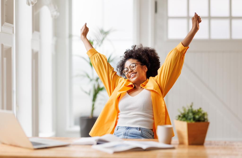 Joyful African American business woman freelancer celebrating with arms up after finishing a project on her laptop in home office