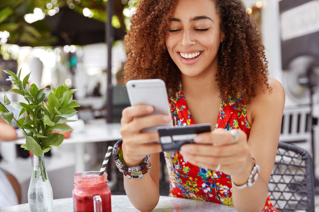 Cheerful young dark-skinned female holding a smartphone and credit card, engaged in online banking or shopping in a cafe interior