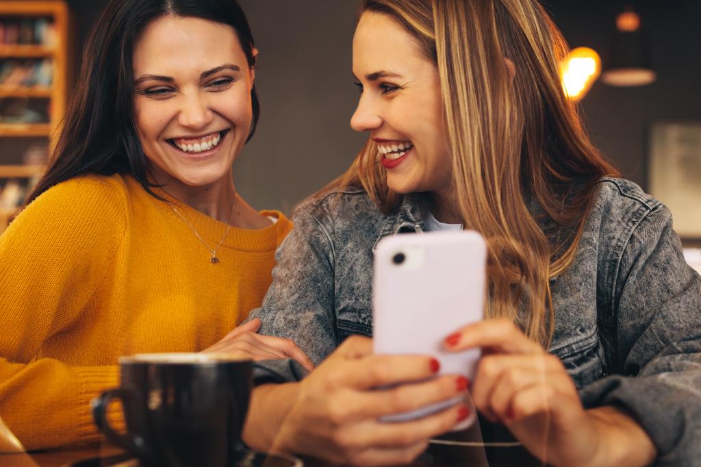 Cheerful young women sitting together at a coffee shop, looking at a mobile phone