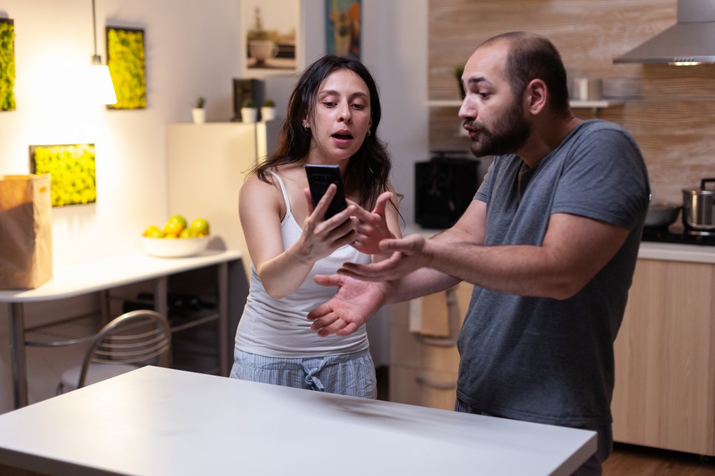 man and a woman arguing in a kitchen over secret messages on his smartphone, representing a marriage crisis due to infidelity and trust issues