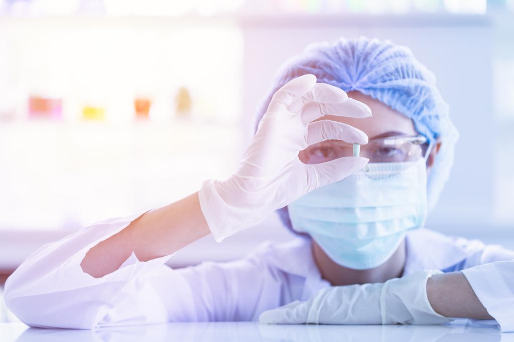 Close-up of an Asian female scientist wearing gloves and mask, holding a small medicine capsule with blurred colorful test tubes and beakers in the background.