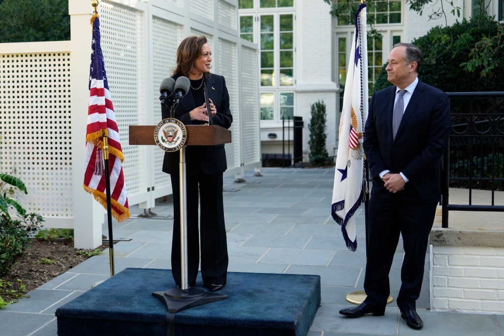 Vice President Kamala Harris speaking at a podium with microphones and flags as Doug Emhoff looks on during a memorial tree planting ceremony, while protesters heckle from outside the gates.