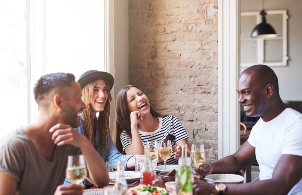 Four diverse friends enjoying dinner and wine together in a restaurant with a brick wall and large window in the background
