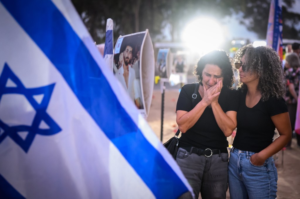 Family and friends gathering at the Nova Festival site in Re'im, Israel to commemorate the anniversary of Hamas October 7 attacks, with two women standing next to a flag