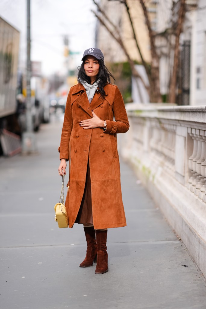 A guest at New York Fashion Week 2024, wearing a NY cap hat, a gray hoodie sweater, a brown-orange suede trench coat, and dark suede boots, holding a yellow leather bag