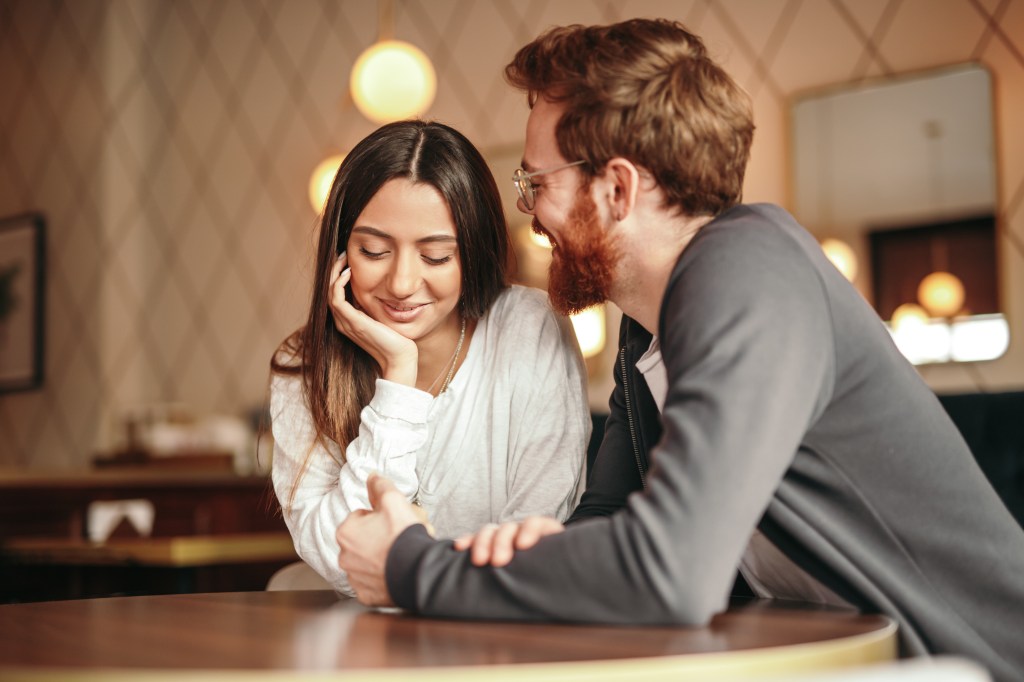 Aaron Ashmore and young woman, both in stylish attire, happily conversing and smiling at a cafe table.