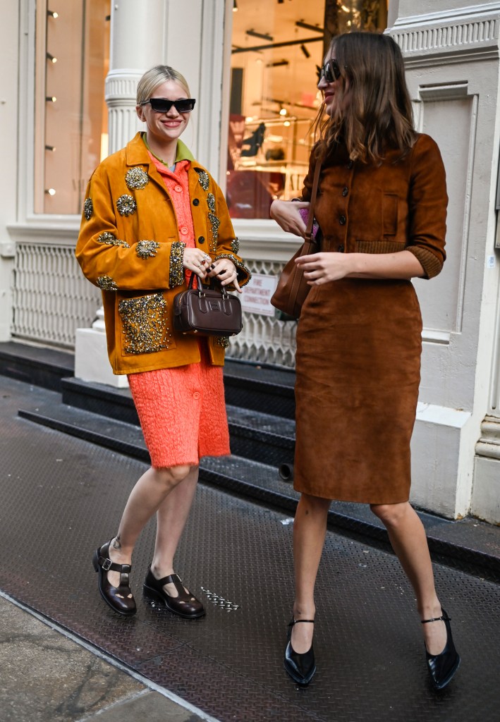 Two models at New York Fashion Week 2024; one wearing an orange and gold embellished jacket, orange Miu Miu dress with a brown bag and black shoes, and the other in a brown Miu Miu dress with black shoes.