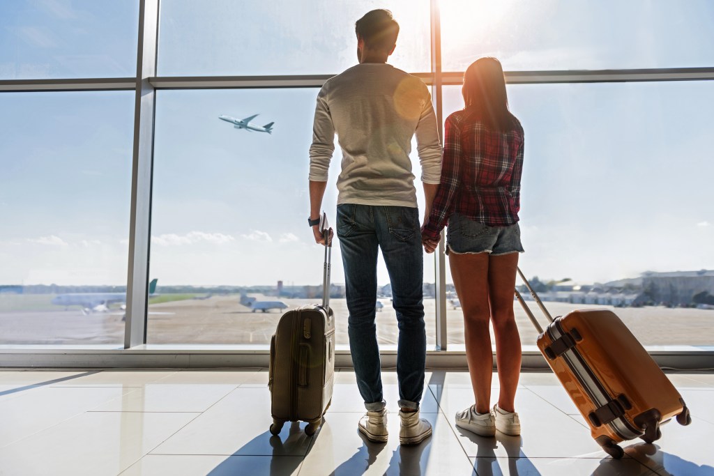 Young man and woman holding hands and looking at a flying plane through airport window, while carrying luggage